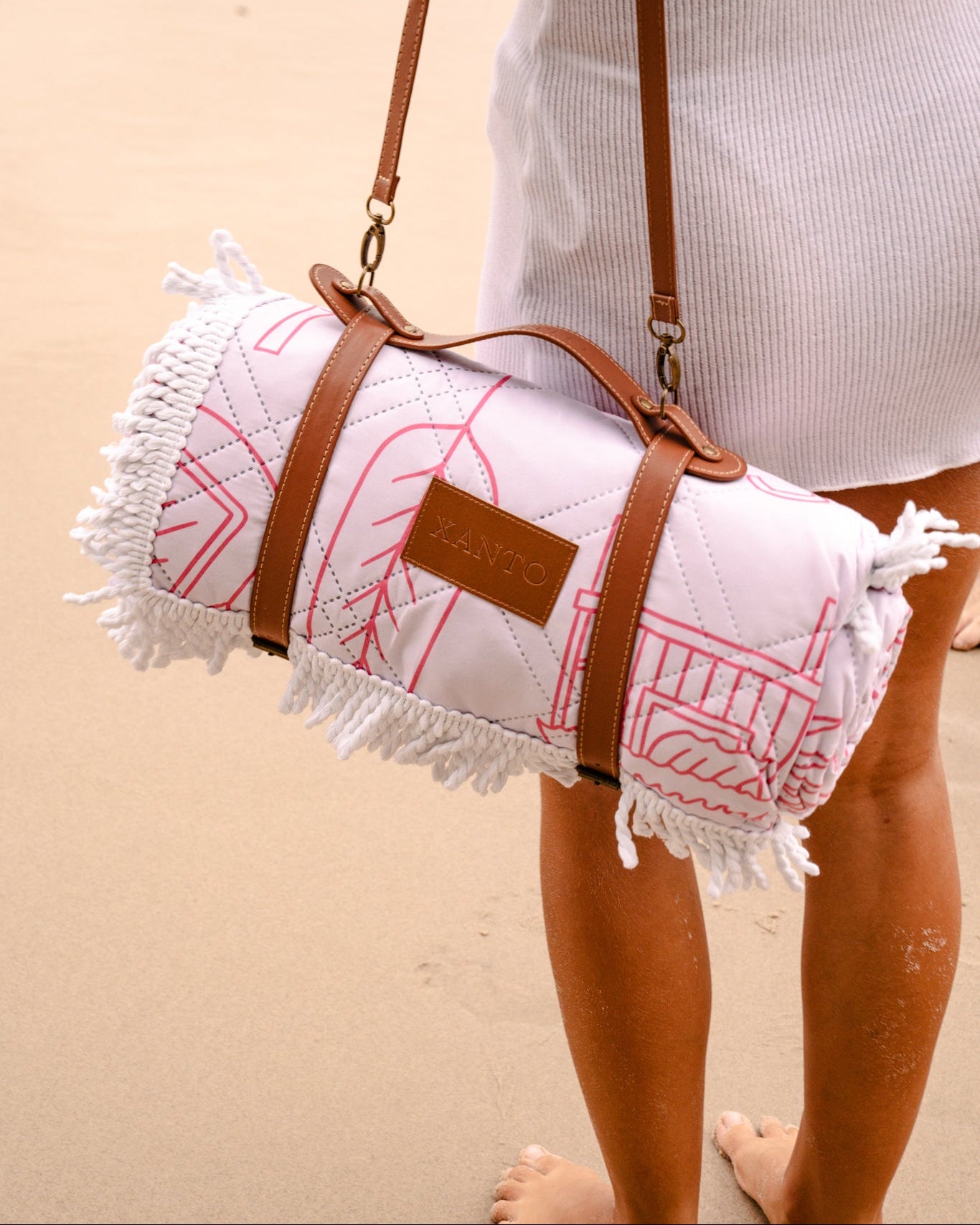 Person holding a rolled-up pink beach towel with a brown strap on a sandy background