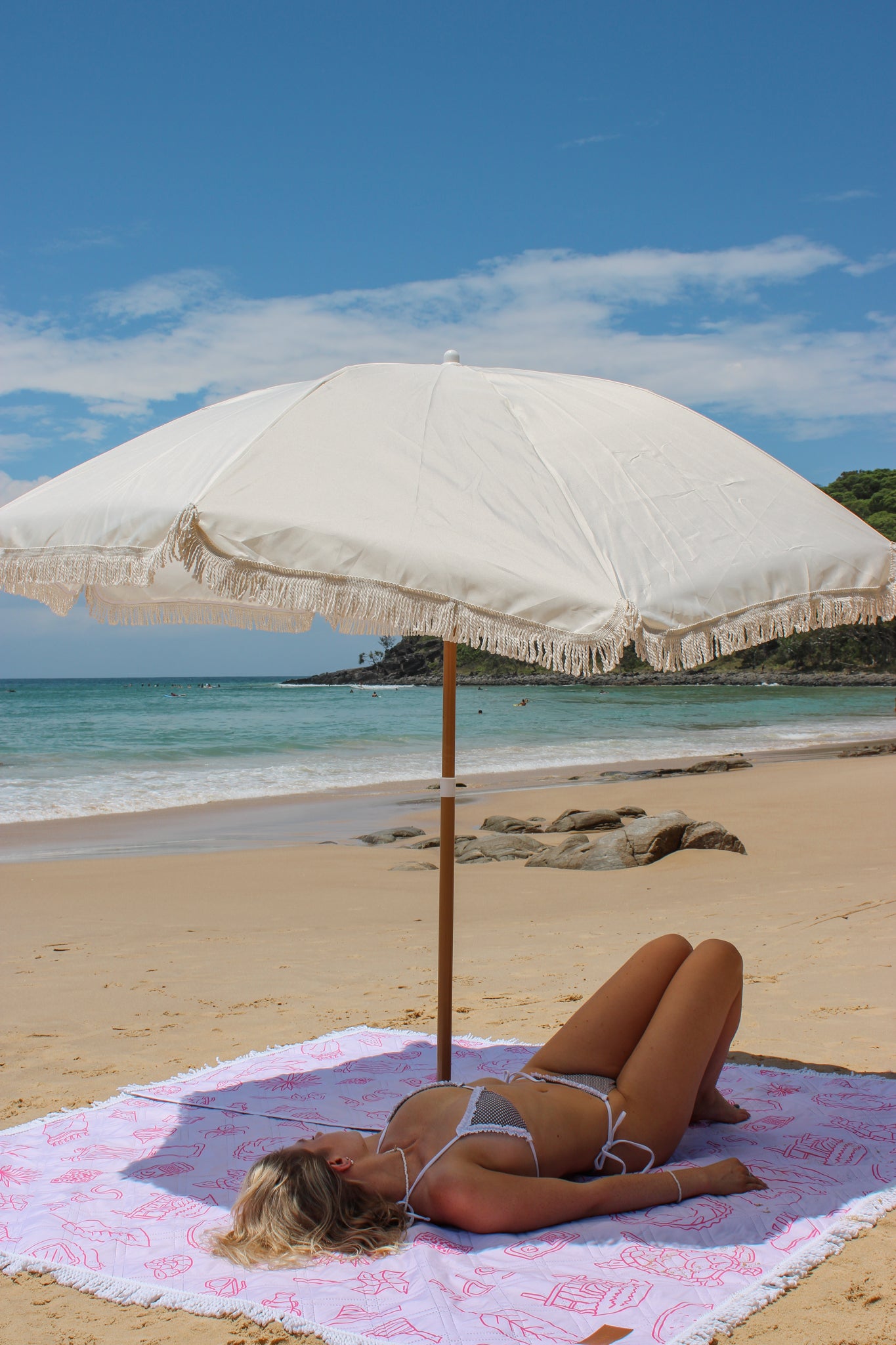 Woman lying on a towel under a large beach umbrella on a sandy beach with ocean and sky in the background.